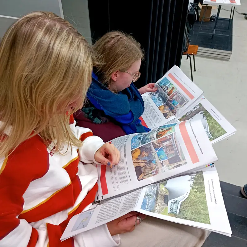 Twee personen lezen een gepersonaliseerd onderzoeksmagazine met foto's, artikels en projectresultaten tijdens een les- of evaluatiemoment.