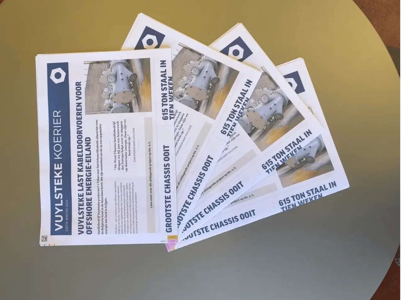 Meerdere exemplaren van een gepersonaliseerde bedrijfskrant van Vuylsteke liggen klaar voor verspreiding en tonen gerealiseerde staalbouwprojecten en bedrijfsnieuws.