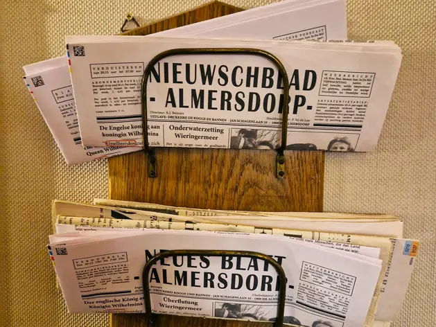 Houten krantenrek gevuld met meerdere exemplaren van een lokale dorpskrant, klaar om meegenomen en gelezen te worden.