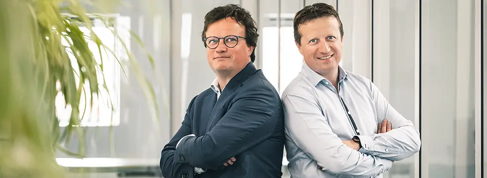 Twee zaakvoerders staan rug aan rug in een moderne kantooromgeving en vertegenwoordigen het leiderschap achter hun organisatie.